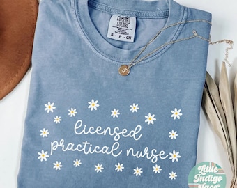May include: A blue jean t-shirt with the text "licensed practical nurse" in white cursive lettering surrounded by white daisy flowers. The shirt has a tag that reads "Comfort Colors" and "S-P-CH".