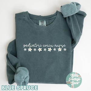 May include: A teal blue sweatshirt with the text "pediatric cvicu nurse" in white lettering and a row of white daisies with yellow centres. The sweatshirt is folded in half and has a small tag with the text "Little Indigo Lace" in the bottom right corner.