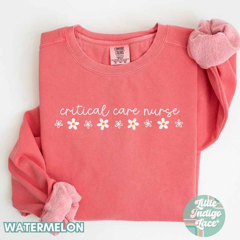 May include: A pink sweatshirt with the text "critical care nurse" in white lettering and a row of white daisies below the text. The sweatshirt is folded in half and the sleeves are rolled up.