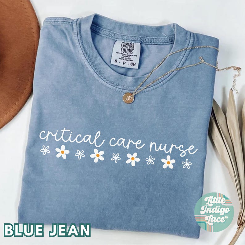 Puede incluir: Una camiseta azul jean con el texto "critical care nurse" y una fila de margaritas blancas con centros amarillos. La camiseta est&aacute; hecha de un material suave y c&oacute;modo y tiene un corte holgado.