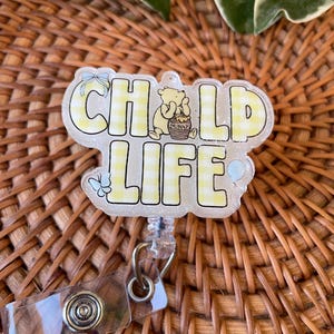 May include: A clear badge reel with the words "CHILD LIFE" in yellow checkered letters. A cartoon bear is holding a honey pot. The badge reel has a clear plastic strap and a metal clip.