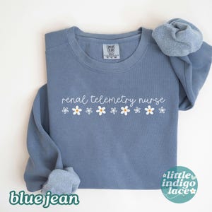 May include: A blue jean colored sweatshirt with the words "renal telemetry nurse" in white script, with small white and yellow daisy graphics. The sweatshirt has a crew neck and long sleeves. The words "blue jean" are at the bottom left.