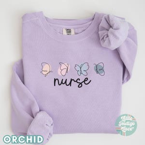 May include: A lavender sweatshirt with the word "nurse" and a line drawing of four butterflies in different colors. The butterflies are pink, blue, and light blue.
