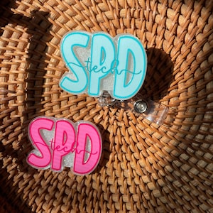 May include: Two acrylic badge reels featuring the text SPD tech in a bold, rounded font. One badge reel is light blue with a glittery background, and the other is bright pink with a glittery background. Both have a clear plastic clip attachment.