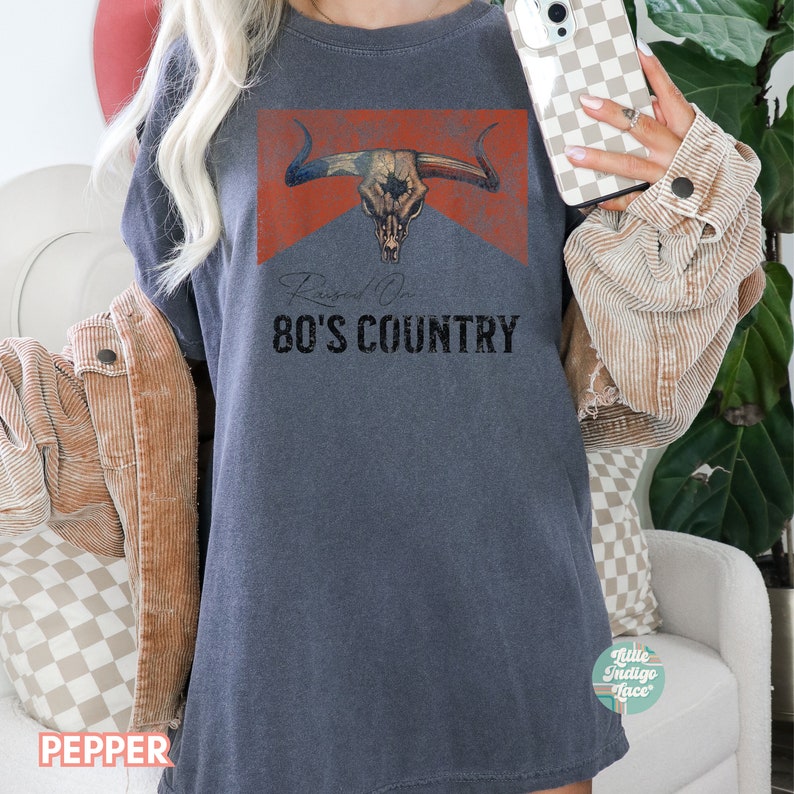 May include: A dark gray t-shirt with a distressed red, white, and blue graphic of a bull skull with the text "Raised On 80's Country".