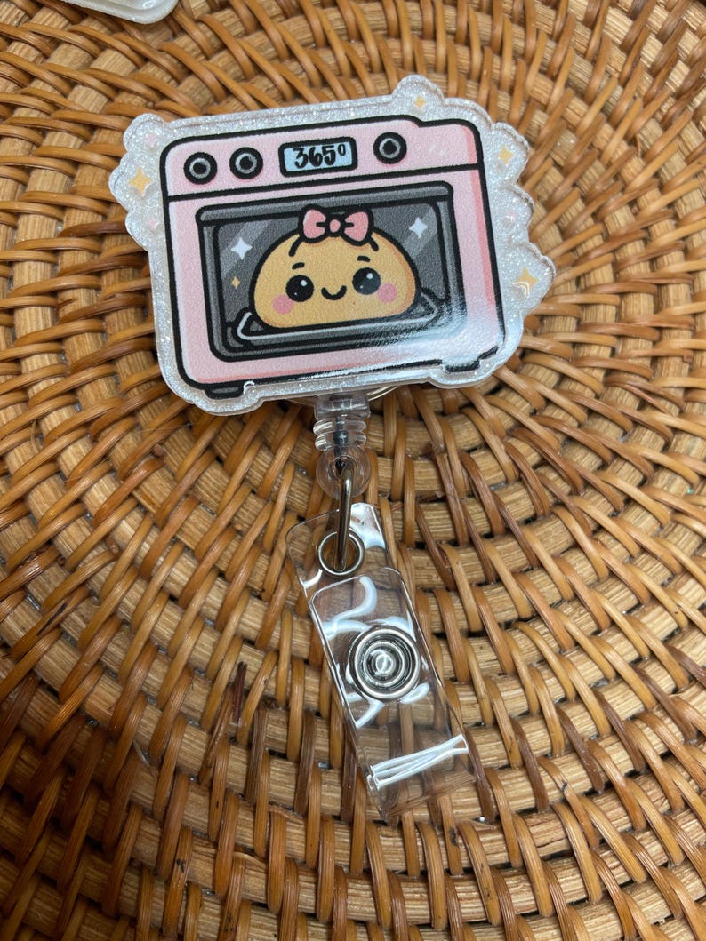 K&ouml;nnte beinhalten: Ein rosa-wei&szlig;es Abzeichen mit Ofenform und einem Cartoon-Kn&ouml;del im Inneren. Der Ofen zeigt die Zahl "365&deg;". Das Abzeichen hat eine transparente, einziehbare Kordel und einen Metallclip.