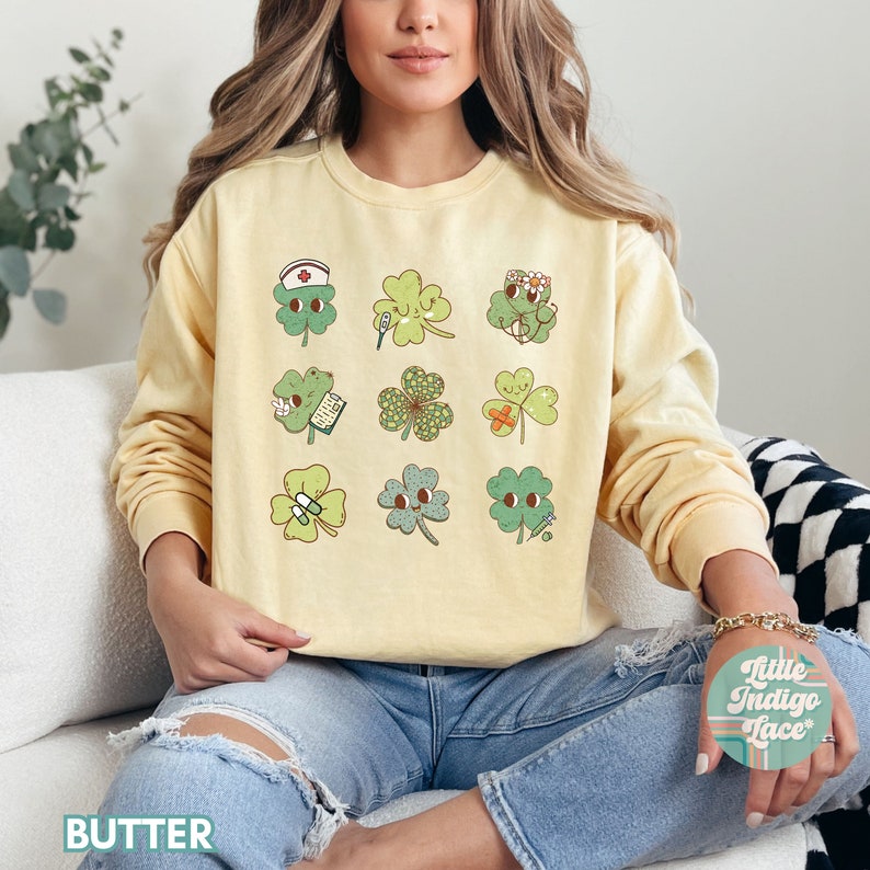 K&ouml;nnte beinhalten: Ein gelber Pullover mit einem Aufdruck von 12 gr&uuml;nen vierbl&auml;ttrigen Kleebl&auml;ttern, jedes mit einem anderen niedlichen Gesicht und einem kleinen Detail wie einem Verband, einer Pille oder einem Stethoskop. Der Pullover wird von einer Person getragen, die auf einem wei&szlig;en Sofa sitzt.