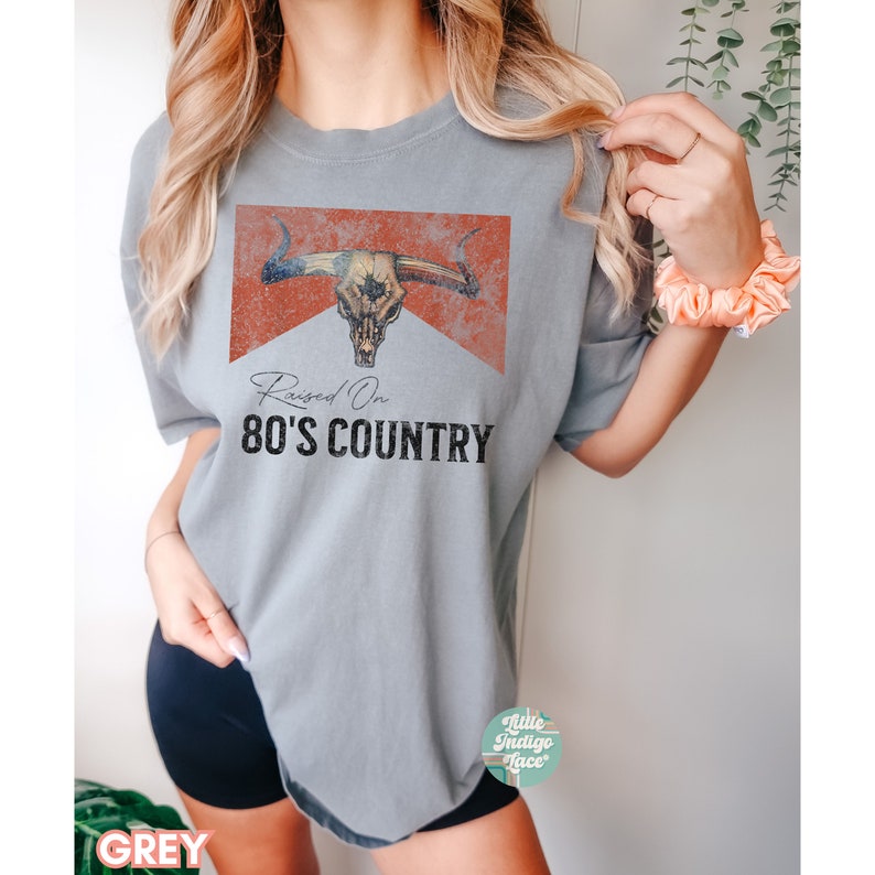 May include: A grey t-shirt with a distressed red and white bull skull graphic and the text "Raised On 80's Country".