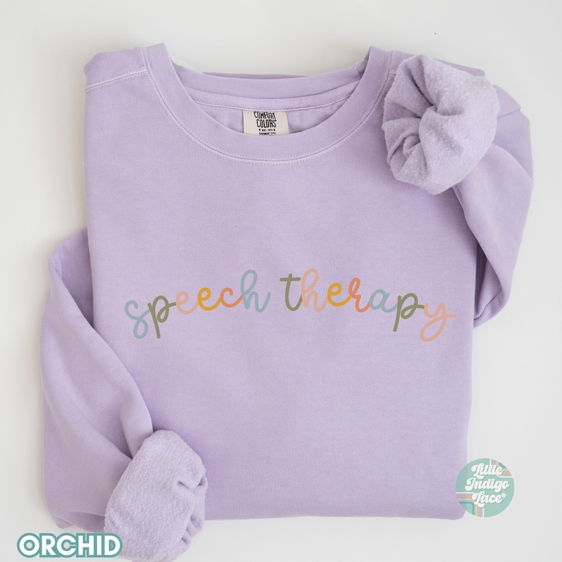 May include: A lavender sweatshirt with the text "speech therapy" in colorful lettering. The sweatshirt is folded and has a small tag on the collar.