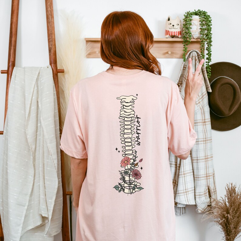 Puede incluir: Camiseta rosa con un dise&ntilde;o floral y una ilustraci&oacute;n estilizada de una columna vertebral humana con la palabra "support" escrita verticalmente a lo largo de la columna vertebral.
