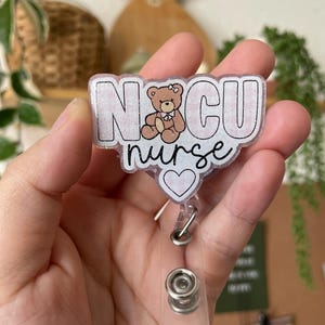 May include: A pink and white glitter badge holder with the text "NCU nurse" and a small cartoon bear with a pink bow. The badge holder has a clear plastic retractable clip.