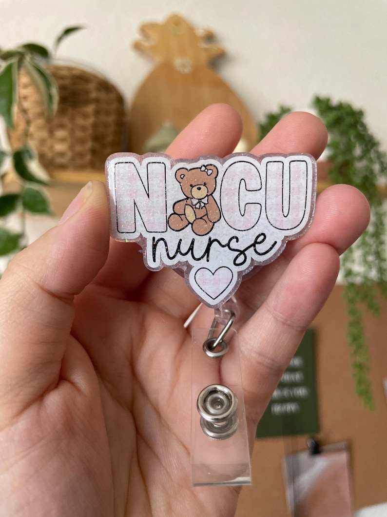 May include: A pink and white glitter badge holder with the text "NCU nurse" and a small cartoon bear with a pink bow. The badge holder has a clear plastic retractable clip.
