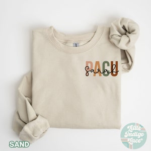 May include: A beige sweatshirt with the text "PACU" in colorful letters and the word "Sarah" written in cursive underneath. The sweatshirt is folded in half and the text is centered on the front of the sweatshirt.