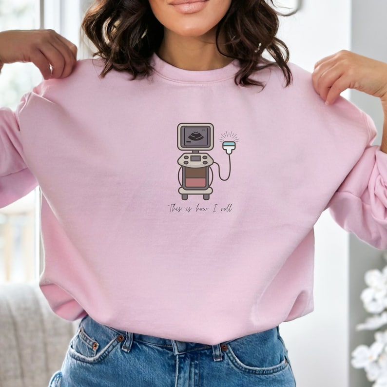 May include: Light pink sweatshirt featuring a medical ultrasound machine graphic. The illustration includes a screen, controls, and a probe, with the text "This is how I roll" below. The design is in shades of brown, gray, and white.