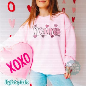 May include: Light pink sweatshirt with the word "neur" in a checkered pattern and a brain graphic. The sweatshirt has several heart-shaped candies with phrases like "I love you" and "XOXO". A pink heart-shaped balloon with "XOXO" is also visible.