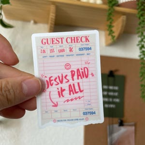 May include: A pink and white sticker with a receipt design. The sticker says "JESUS PAID it ALL" in red lettering. The sticker has a check mark and a smiley face.