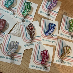 Pu&ograve; includere: Assortimento di porta badge retrattili pieni di glitter colorati con un design a gamba di scheletro bianco. Ogni porta badge &egrave; attaccato a una carta con il testo "Little Indigo Lace".