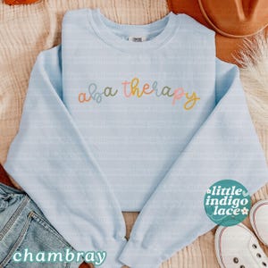 K&ouml;nnte beinhalten: Hellblaues Sweatshirt mit Rundhalsausschnitt und dem Schriftzug "aba therapy" in Pastellfarben. Das Sweatshirt wird mit Jeans, einem Hut und Turnschuhen gezeigt. Das Wort "chambray" ist unten links aufgedruckt.