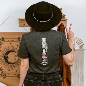 Puede incluir: Una camiseta gris oscuro con un dise&ntilde;o floral y una columna vertebral estilizada con la palabra "apoyo" escrita en ella. La camiseta la lleva puesta una persona que lleva un sombrero negro y pantalones vaqueros.