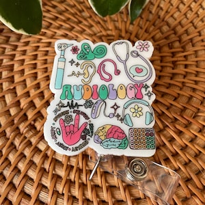 May include: A white badge reel with the word "Audiology" in colorful letters. The badge features various medical-themed illustrations, including a stethoscope, ear, and brain. The reel has a clear plastic clip and a metal retractable cord.