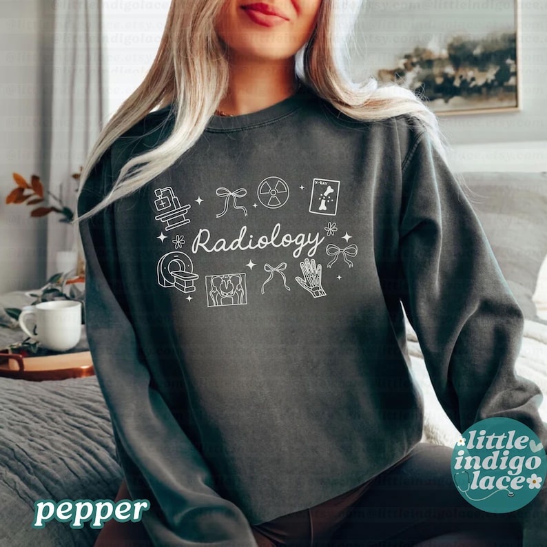 May include: A dark gray sweatshirt with the word "Radiology" in white script, surrounded by white medical-themed graphics. The sweatshirt has a crew neck and long sleeves. The word "pepper" is printed in white at the bottom left.
