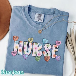 May include: A blue jean colored t-shirt with the word "NURSE" in large, colorful letters. The letters are decorated with heart-shaped candies with phrases like "Love Rx" and "Code Love". A gold necklace rests on the shirt.