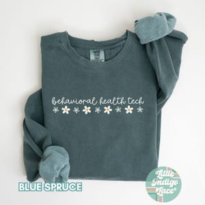 May include: A teal blue sweatshirt with the text "behavioral health tech" in white lettering and a row of white daisies with yellow centers. The sweatshirt is made by "Comfy Colors" and has a "Little Indigo Lace" tag.