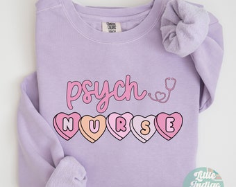 Könnte beinhalten: Ein lavendelfarbener Pullover mit dem Text "psych nurse" in rosa und weißen Buchstaben. Das Wort "nurse" ist mit rosa Herzchen aus Bonbons ausgeschrieben. Neben dem Wort "psych" ist ein Stethoskop gezeichnet.