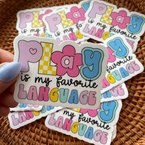 May include: A stack of colorful stickers with the text "Play is my favorite language" in a playful font. The stickers are in various shades of pink, blue, yellow, and green.