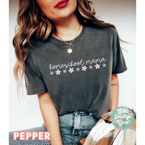 May include: A dark gray t-shirt with the text "homeschool mama" in white script with a row of white daisies underneath. The shirt is being worn by a person with long brown hair.