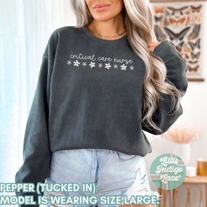 May include: A dark gray sweatshirt with the text "critical care nurse" and a row of white flowers with yellow centers. The model is wearing the sweatshirt tucked in and is wearing blue denim shorts.