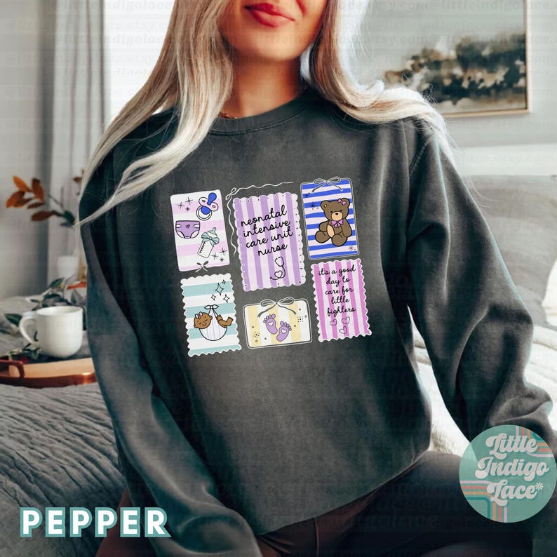 Puede incluir: Sudadera gris oscuro con un dise&ntilde;o gr&aacute;fico colorido que presenta ilustraciones relacionadas con beb&eacute;s y texto. El texto incluye "neonatal intensive care unit nurse" y "It's a good day to care for little fighters."