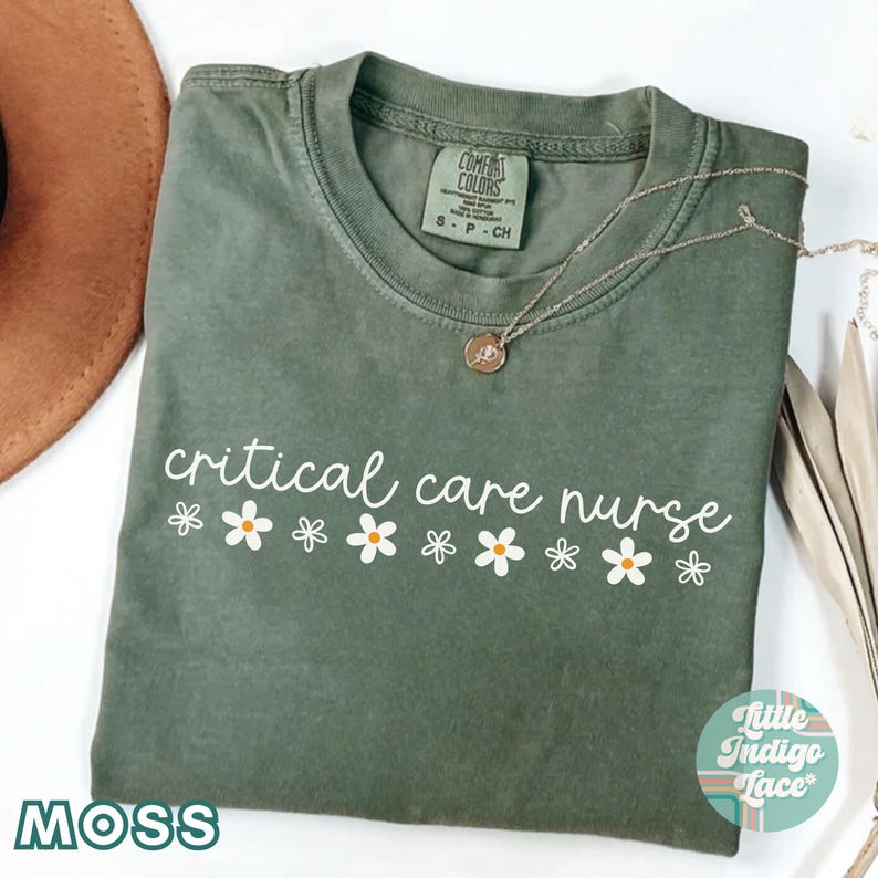 Puede incluir: Una camiseta verde con el texto "critical care nurse" en letras blancas y una fila de margaritas blancas con centros amarillos. La camiseta est&aacute; doblada y tiene una etiqueta que dice "Comfort Colors" y "S-P-CH".