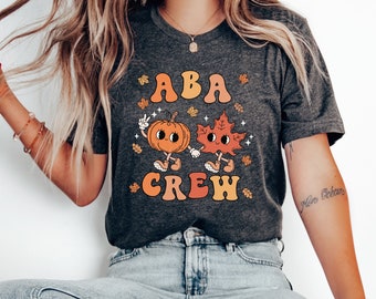 May include: Dark grey t-shirt with the text "ABA CREW" in orange and yellow. The shirt features a cartoon pumpkin and leaf holding hands. The pumpkin is giving a peace sign. Autumn-themed clothing.