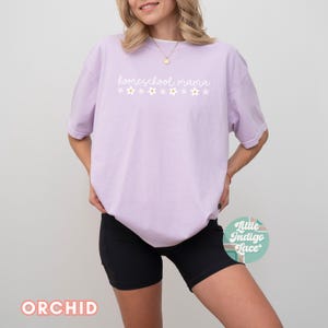 May include: A woman wearing a light purple t-shirt with the text "homeschool mama" and white flowers printed on it. She is also wearing black biker shorts.