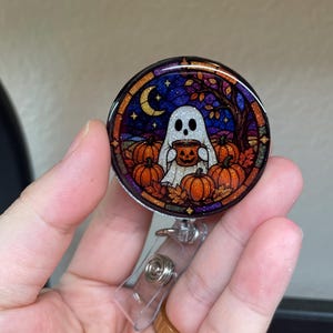 May include: A circular badge reel featuring a stained-glass style illustration of a white ghost holding a jack-o'-lantern mug. The scene includes orange pumpkins, autumn leaves, a crescent moon, and a dark blue and purple starry night sky.