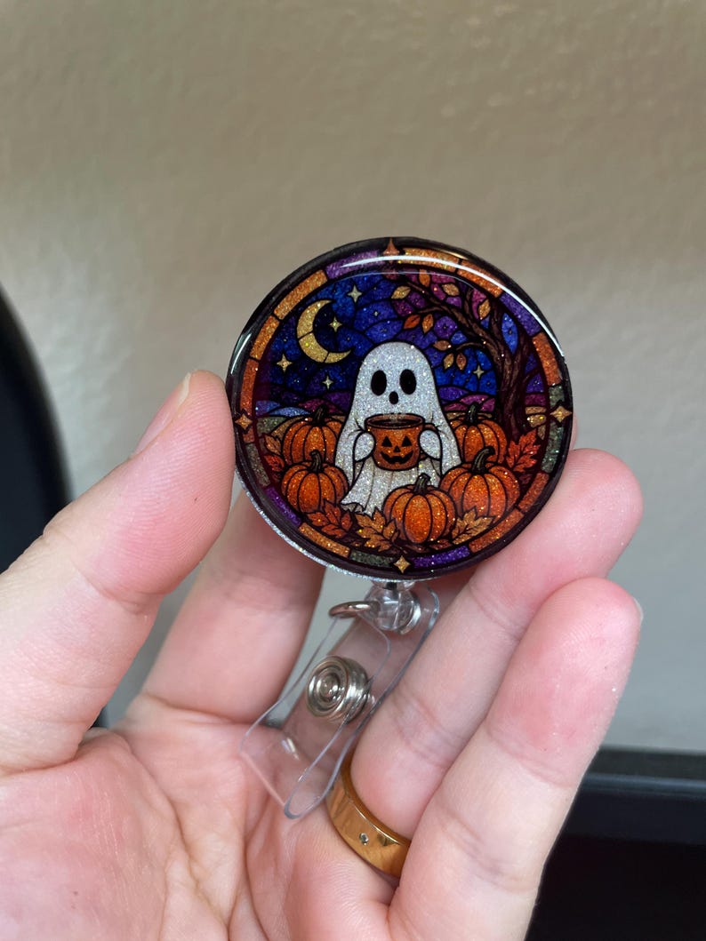 May include: A circular badge reel featuring a stained-glass style illustration of a white ghost holding a jack-o'-lantern mug. The scene includes orange pumpkins, autumn leaves, a crescent moon, and a dark blue and purple starry night sky.
