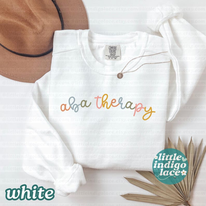 K&ouml;nnte beinhalten: Wei&szlig;es Sweatshirt mit dem Text "aba therapy" in Pastellfarben. Das Sweatshirt hat lange &Auml;rmel und einen Rundhalsausschnitt. Ein brauner Hut und eine goldene Halskette sind im Hintergrund zu sehen. Das Wort "white" steht unten.