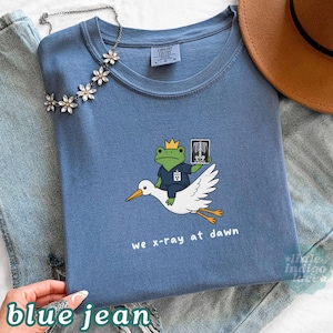 May include: A blue crew neck t-shirt featuring a graphic of a green frog wearing a yellow crown and blue scrubs, riding a white stork while holding an x-ray film. The text below the graphic reads, we x-ray at dawn. The shirt has a Comfort Colors label.