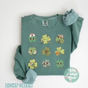 K&ouml;nnte beinhalten: Hellgr&uuml;ner Pullover mit einem Motiv aus neun vierbl&auml;ttrigen Kleebl&auml;ttern, jedes mit einem anderen medizinischen Design. Die Kleebl&auml;tter haben Gesichter und halten Gegenst&auml;nde wie ein Thermometer, einen Verband und eine Pillenflasche. Der Text "Little Indigo Lace" befindet sich in der rechten unteren Ecke des Pullovers.