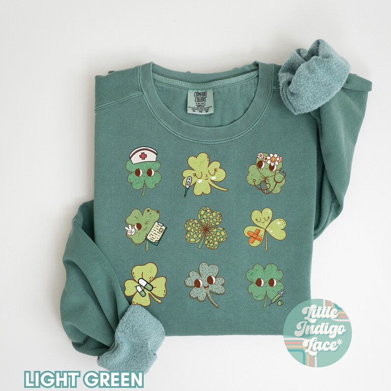 K&ouml;nnte beinhalten: Hellgr&uuml;ner Pullover mit einem Motiv aus neun vierbl&auml;ttrigen Kleebl&auml;ttern, jedes mit einem anderen medizinischen Design. Die Kleebl&auml;tter haben Gesichter und halten Gegenst&auml;nde wie ein Thermometer, einen Verband und eine Pillenflasche. Der Text "Little Indigo Lace" befindet sich in der rechten unteren Ecke des Pullovers.
