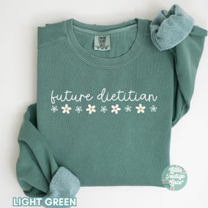 May include: Light green sweatshirt with the words "future dietitian" in white script, decorated with small daisy graphics. The sweatshirt has a crew neck and long sleeves. The words "Light Green" are at the bottom of the image.