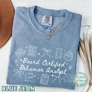 May include: Blue jean Comfort Colors t-shirt with white graphic text and illustrations. The shirt features the words "Board Certified Behavior Analyst" and related icons. A gold necklace with a pendant is draped on the shirt.