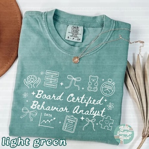 May include: Light green t-shirt with the text "Board Certified Behavior Analyst" and related graphics. The shirt includes the text "Antecedent, Behavior, Consequence" and other icons. A silver necklace with a pendant is draped across the shirt.