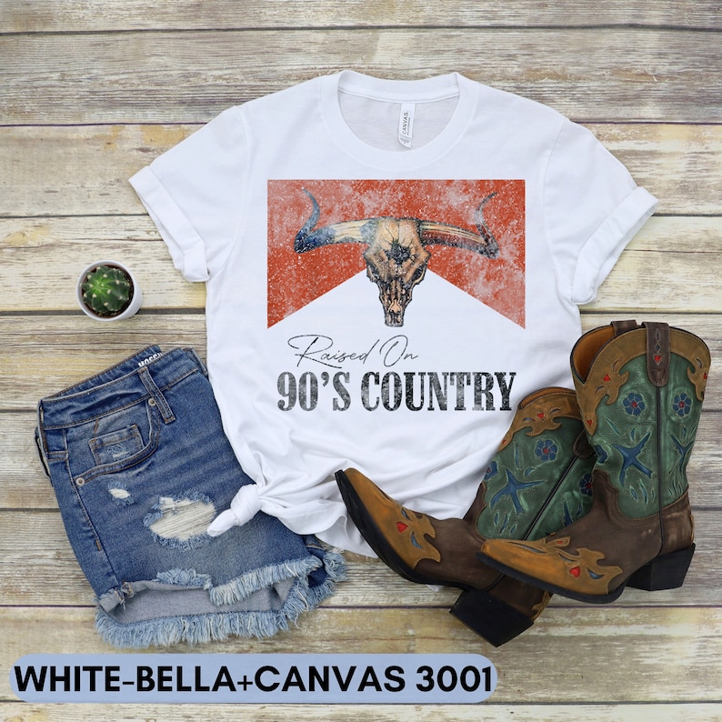 May include: White t-shirt with a red and white graphic of a bull skull with the text "Raised On 90's Country".