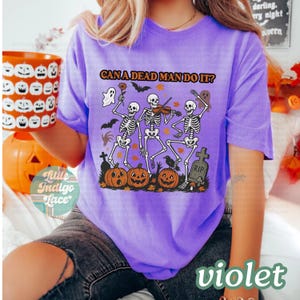 May include: Violet t-shirt with a Halloween-themed graphic. The design features three skeletons playing instruments, pumpkins, bats, and the text "CAN A DEAD MAN DO IT?". A ghost and a tombstone are also included in the design.