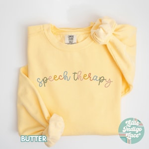May include: A yellow sweatshirt with the words "speech therapy" written in colorful script across the front. The sweatshirt is folded and laid flat on a white surface.
