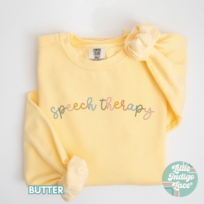 May include: A yellow sweatshirt with the words "speech therapy" written in colorful script across the front. The sweatshirt is folded and laid flat on a white surface.