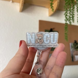 May include: A badge reel with the letters "NCU" in a blue gingham pattern, featuring a teddy bear illustration. The text "neonatal intensive care unit" is below the letters. The reel has a clear plastic strap and a metal clip.