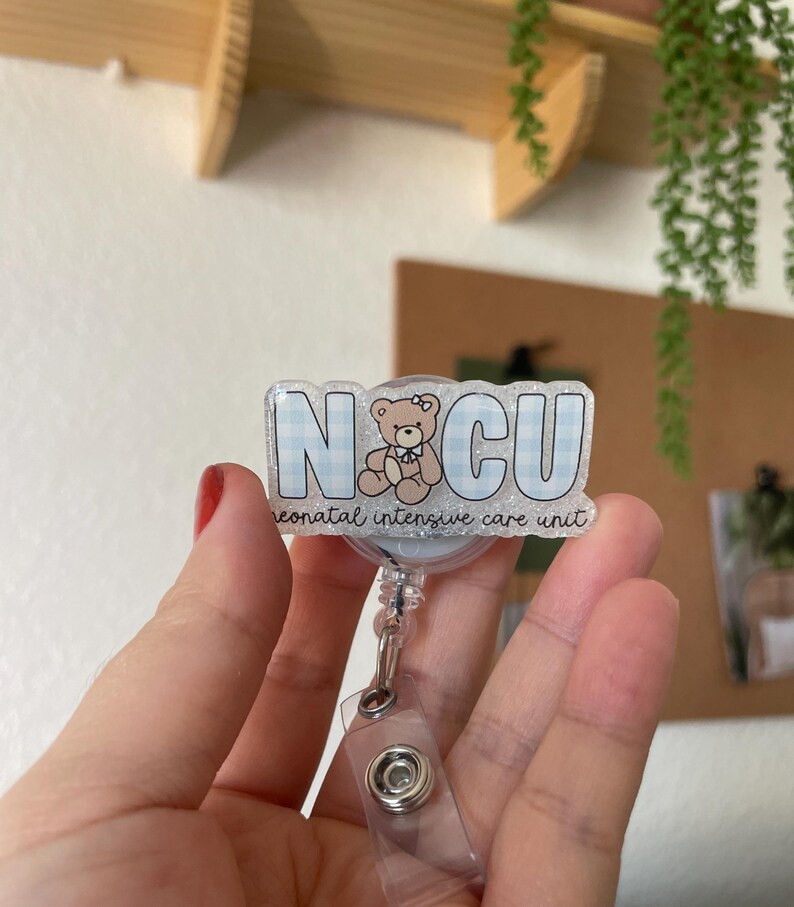 May include: A badge reel with the letters "NCU" in a blue gingham pattern, featuring a teddy bear illustration. The text "neonatal intensive care unit" is below the letters. The reel has a clear plastic strap and a metal clip.